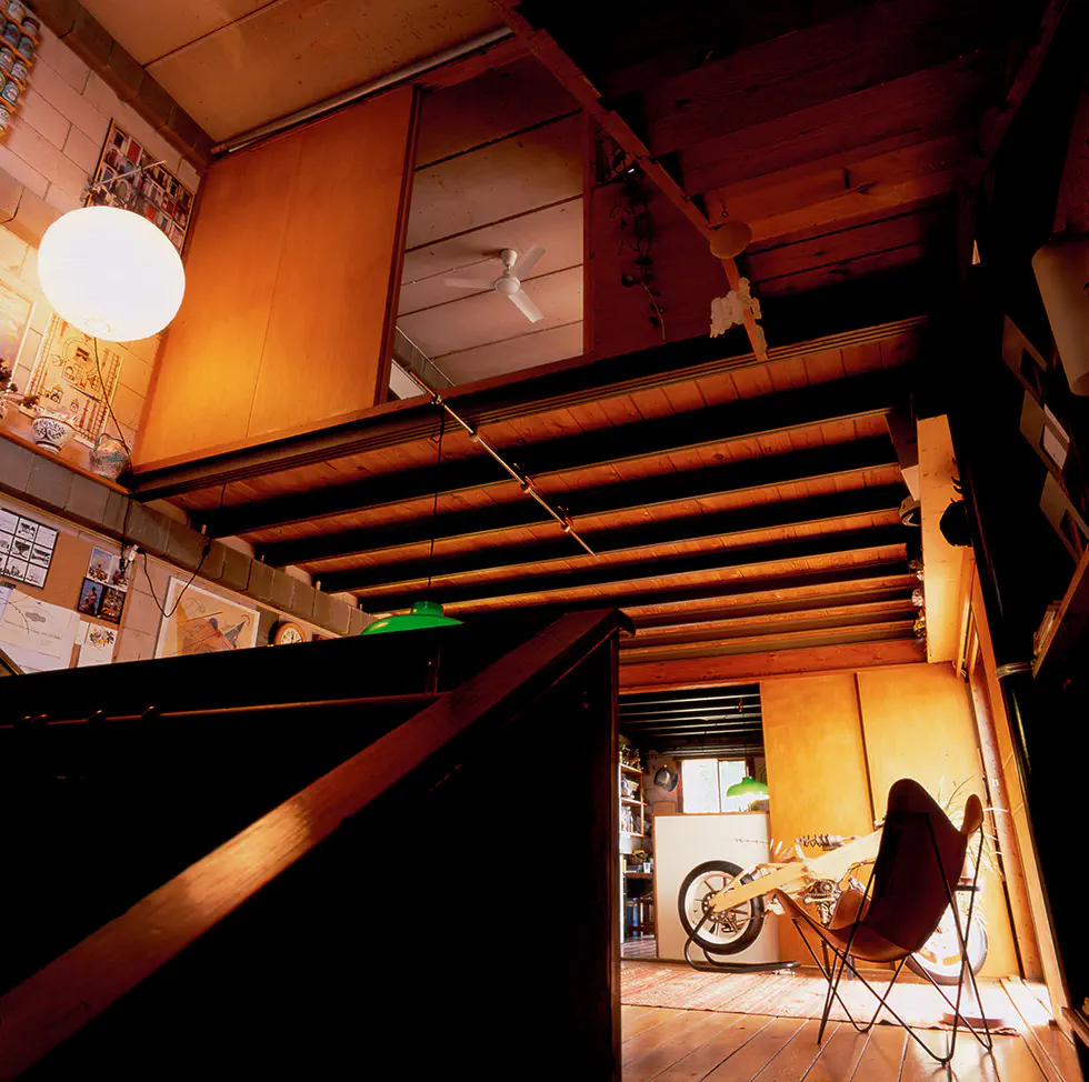 Low-angle interior view of a warm, eclectic space with exposed steel beams, sliding wooden panels opening into a loft area with a ceiling fan. In the ground floor, a wood motorcycle prototype is visible in the background.