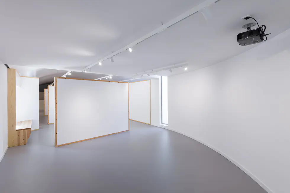 Curved gallery space with modular white walls and wooden frames, a bench built into the wall, overhead track lighting, and a ceiling-mounted projector.