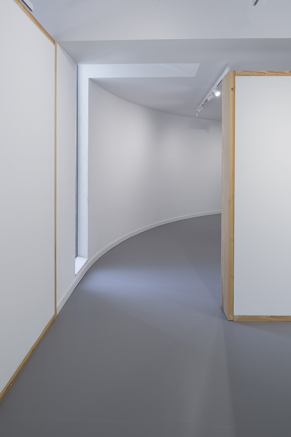 Curved gallery hallway with modular white walls and wooden frames, a tall vertical window providing natural light, and overhead track lighting.