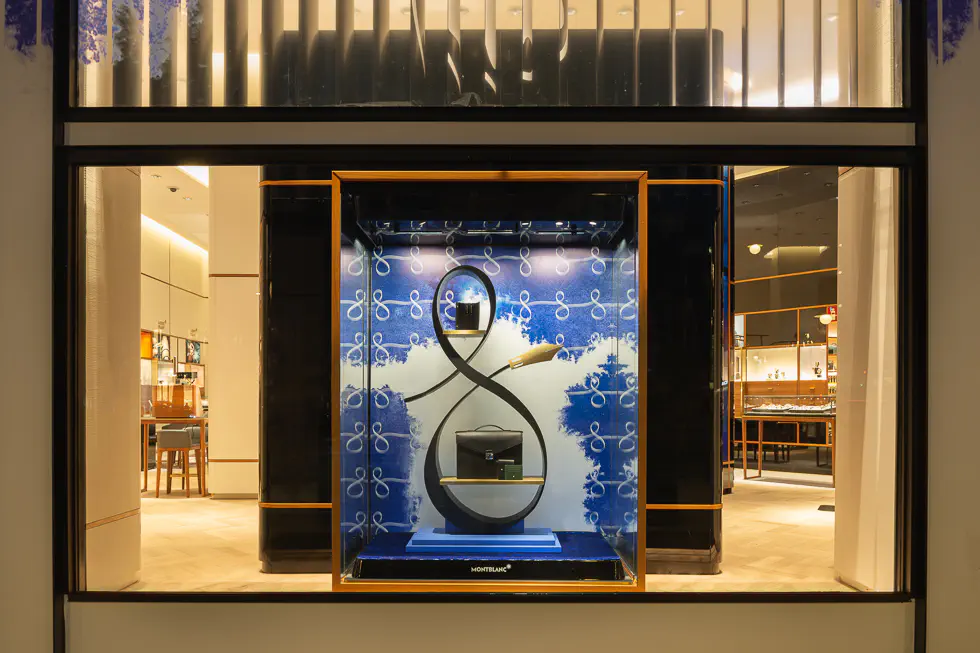 Montblanc store window display featuring a sophisticated arrangement of bags and accessories against a stylish blue background with elegant patterns.