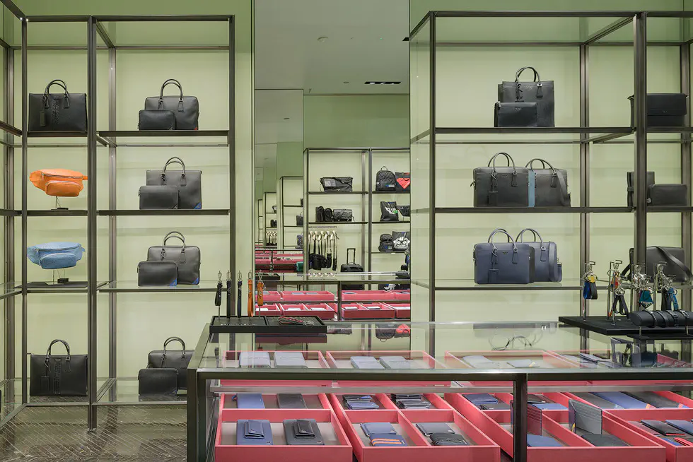 Prada store interior with two facing mirrors featuring an infinity mirror effect, there are shelves of designer bags in various colors, organized neatly against a light green background.