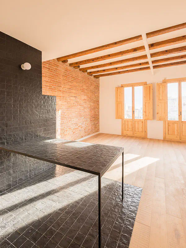 Open-plan living area with black tiled countertop, exposed brick wall, wooden ceiling beams, and natural sunlight coming through two large balconies with wooden shutters.