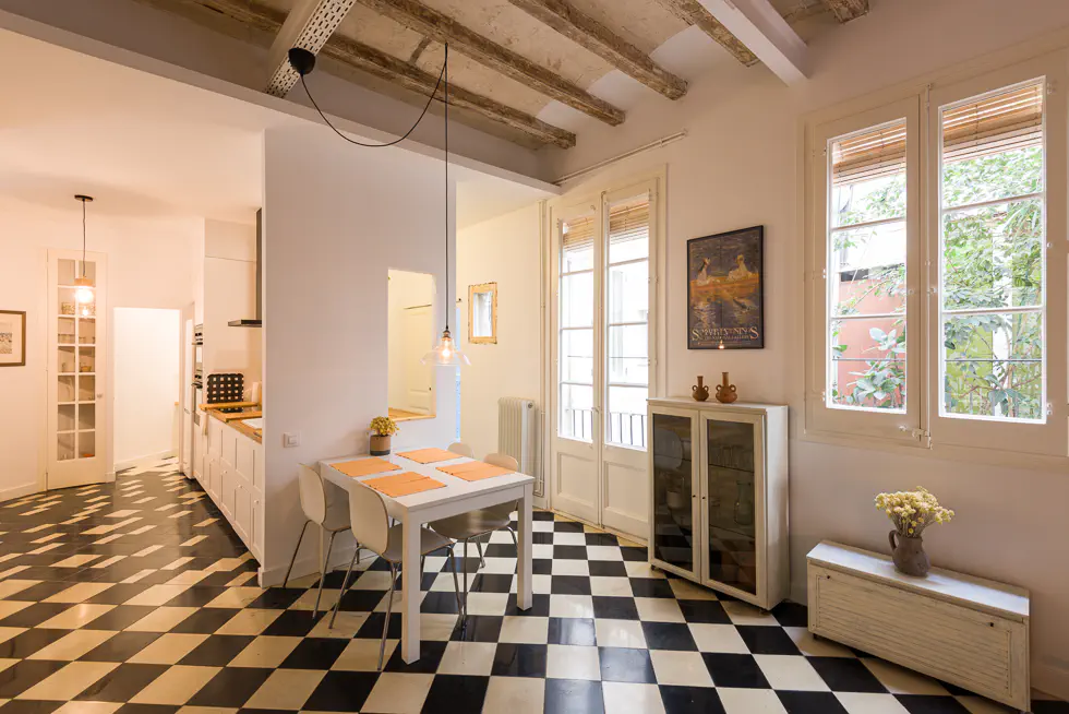 Dining area with checkered floor tiles, exposed beams, a white table with four chairs, and large windows overlooking greenery.