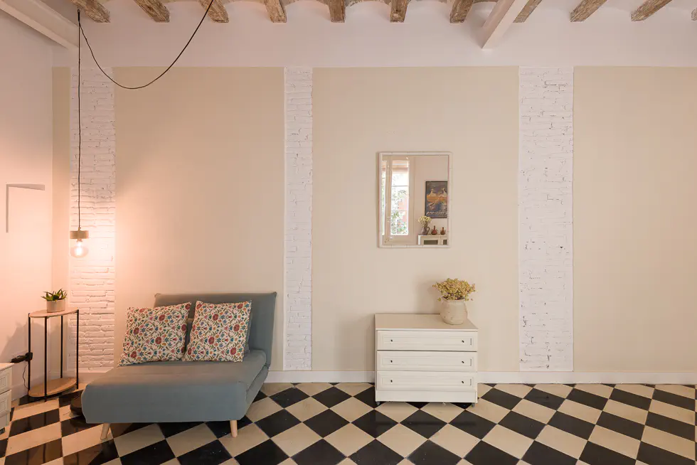 Cozy corner with a blue armchair, floral pillows, a small table, a white dresser, a square mirror on the wall reflecting the door onto the patio and checkered floor tiles.