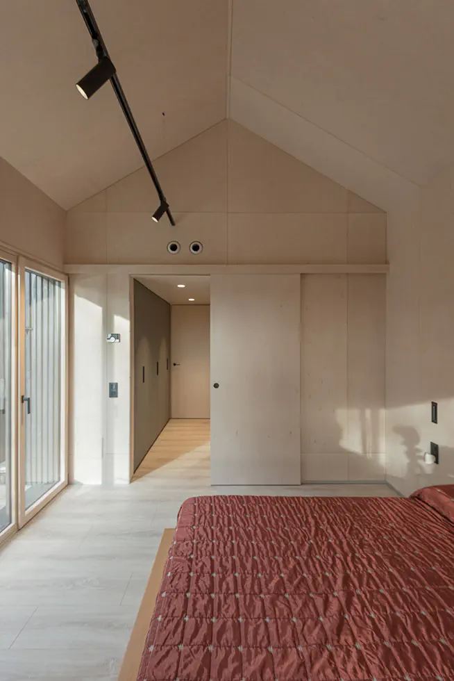 Bedroom with a high pitched ceiling, featuring a red bedspread, large glass doors, and minimalist wooden walls and flooring.
