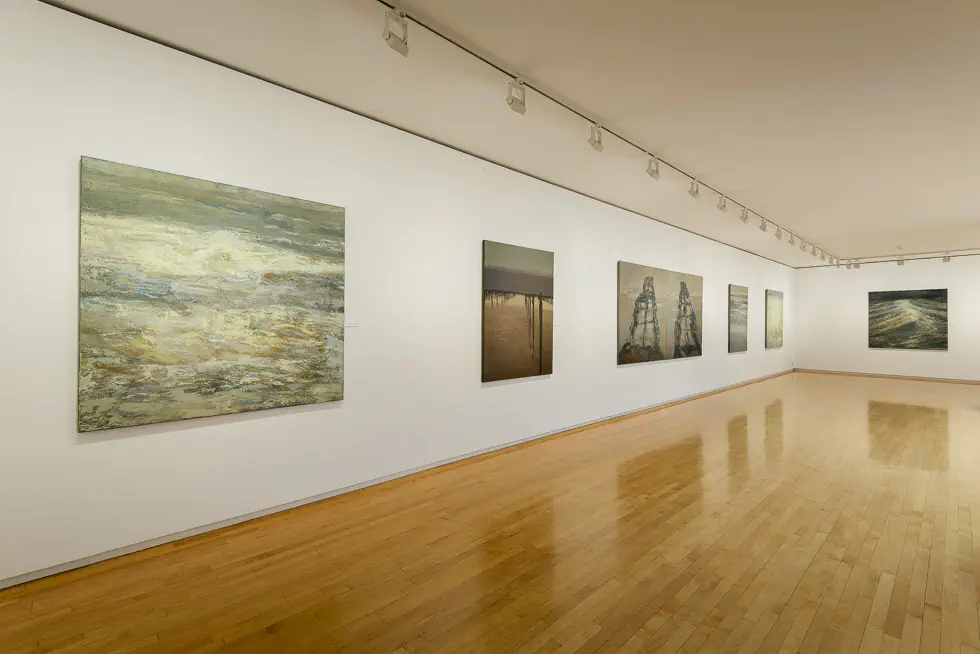 Spacious gallery room with wooden floors displaying large contemporary paintings on white walls, illuminated by track lighting.