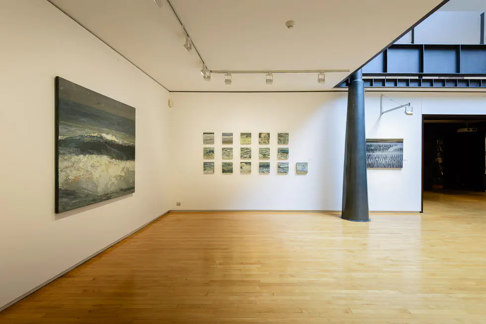 A spacious art gallery with white walls and polished wooden floors, displaying seascape paintings under track lighting. A black conic steel column stands in the side.