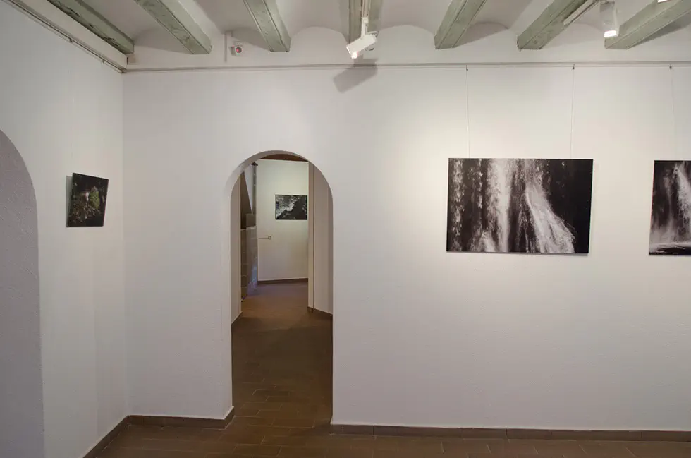 Archway view of gallery with framed photographs on white walls, wooden ceiling beams, and tiled floor.