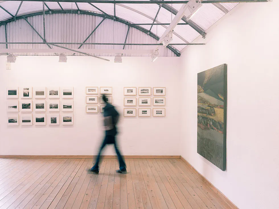 Exhibition view with a person walking past framed artworks.
