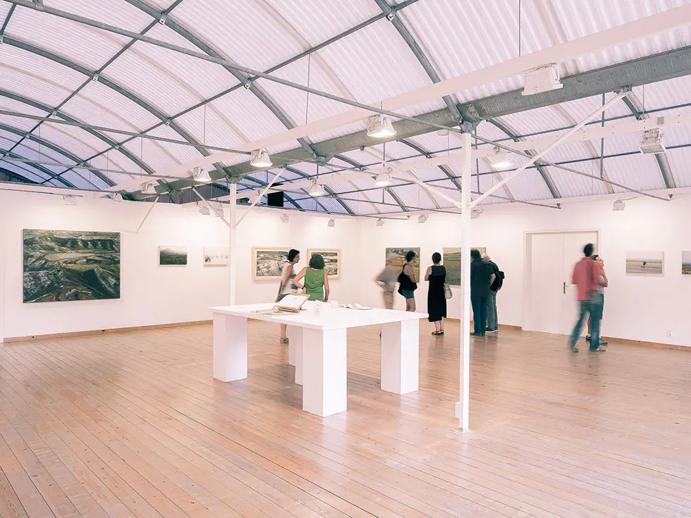 People viewing artworks in a well-lit exhibition space with framed pieces on the walls and a table in the center.