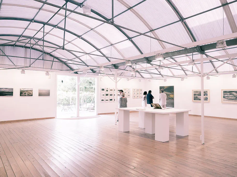 People viewing framed artworks in a spacious, bright exhibition hall with wooden floors and a high, curved ceiling.