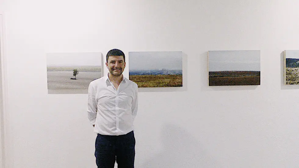 A man in a white shirt standing in front of landscape photographs on a gallery wall. The photographs depict various natural scenes, including fields and distant horizons.
