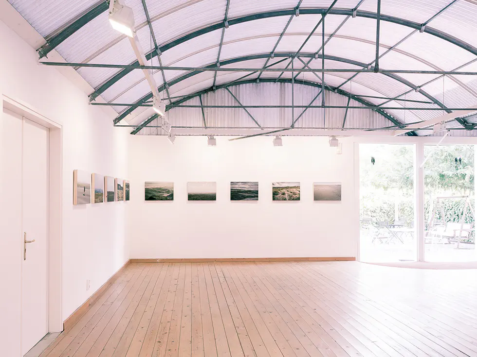 Gallery room with a curved ceiling and landscape photographs on the walls. Large door on the right open to a garden.