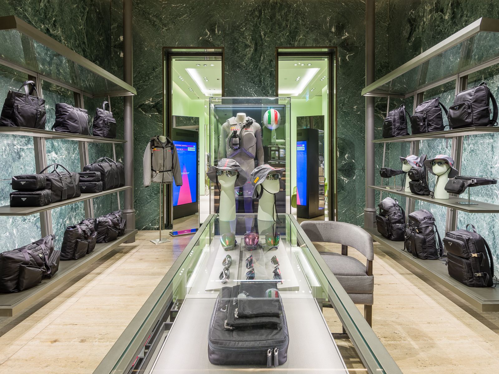 Boutique de luxe Prada amb parets de marbre verd, mostrant sabatilles esportives de dissenyador, bosses negres i accessoris en prestatges de metall elegant. Una taula de vidre mostra ulleres i barrets, mentre que un maniquí amb un vestit de rendiment està tancat en una vitrina de vidre.