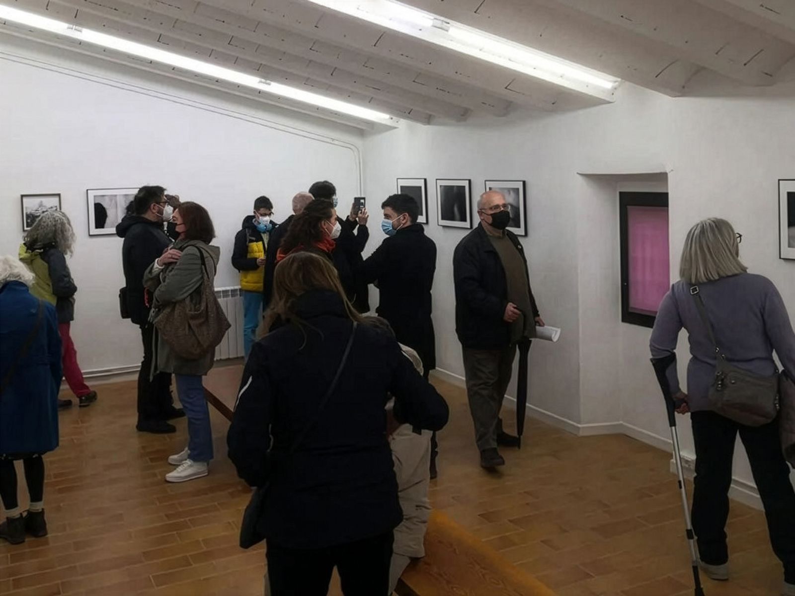 Visitantes viendo fotografías enmarcadas en las paredes de una galería de arte, todos usando mascarillas quirúrgicas y conversando.