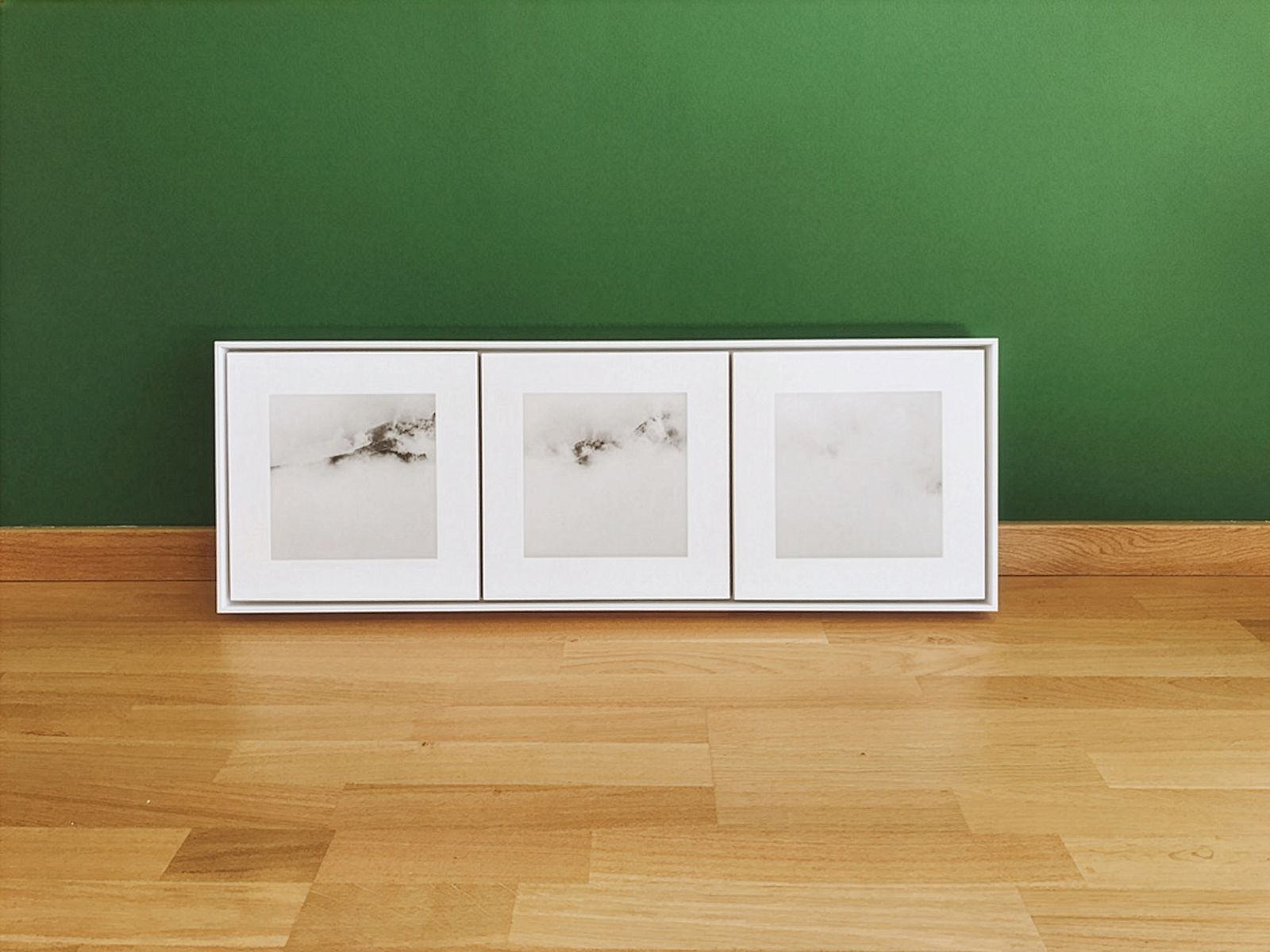 Tríptic d'obra d'art emmarcada amb paisatges muntanyosos coberts de boira exposat contra una paret verda sobre un terra de fusta.