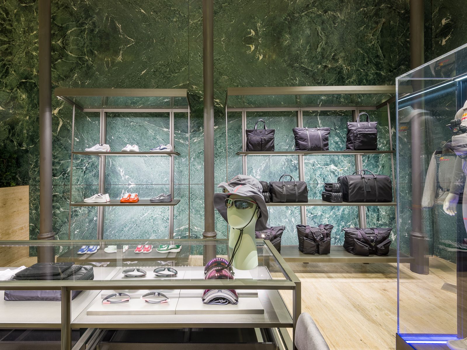 Boutique de luxe amb parets de marbre verd, mostrant sabatilles esportives de dissenyador, bosses negres i accessoris en prestatges de metall elegant. Una taula de vidre mostra ulleres i barrets, mentre que un maniquí amb un vestit de rendiment està tancat en una vitrina de vidre.