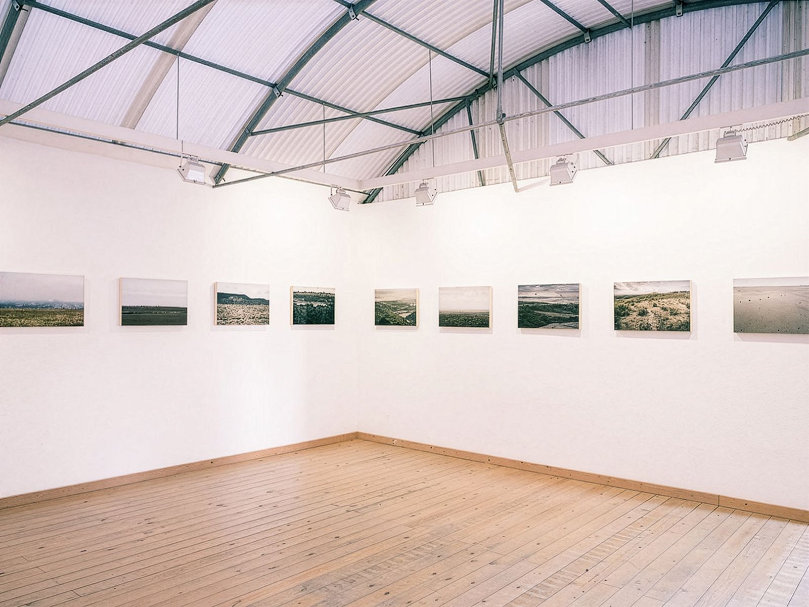 Sala de exposición con una serie de fotografías de paisajes dispuestas en una sola fila a lo largo de las paredes blancas, con un techo curvo alto y suelos de madera.