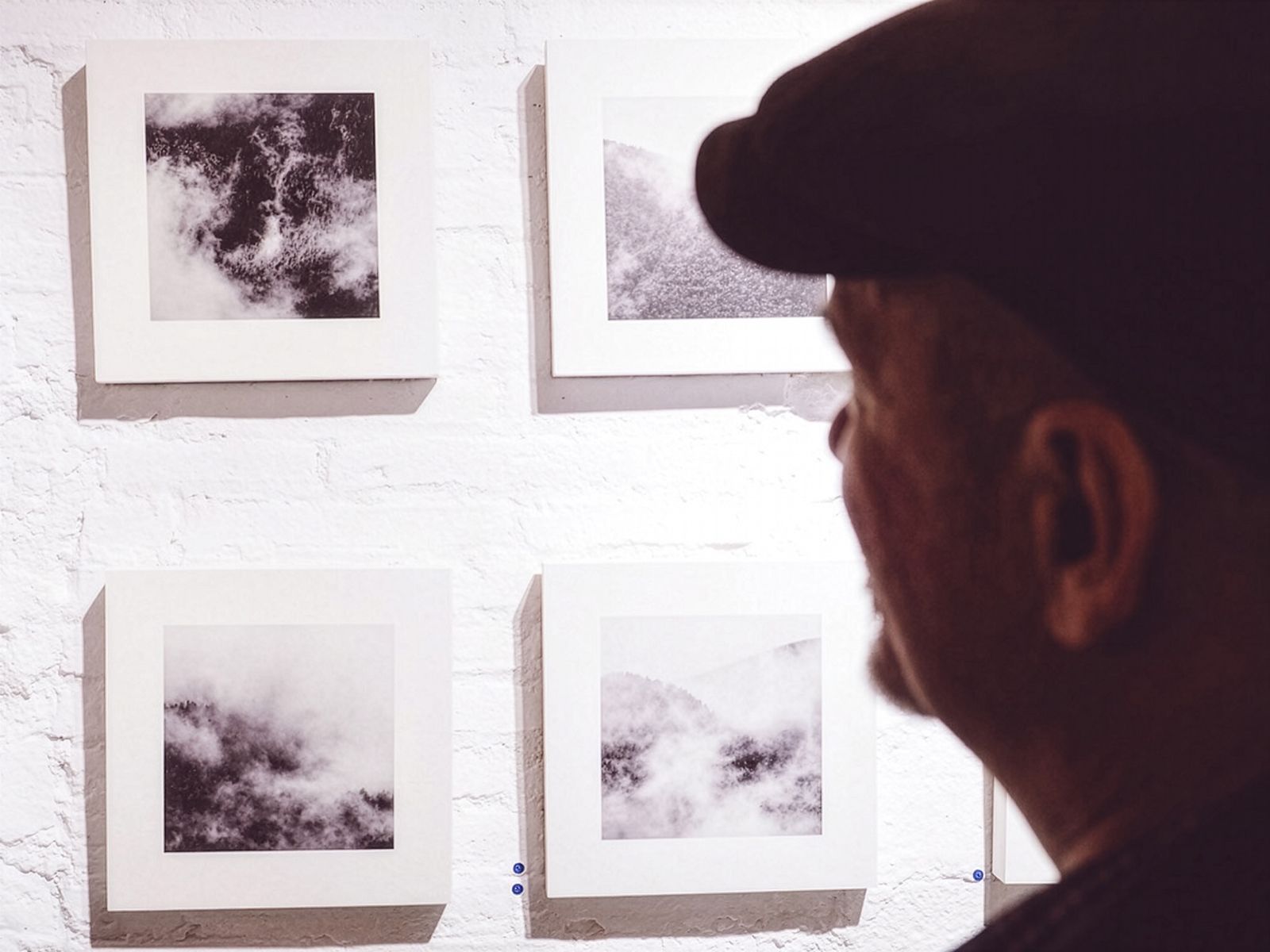 Una persona mirant una sèrie de fotografies quadrades en blanc i negre de núvols i boira en una paret de totxo blanc.