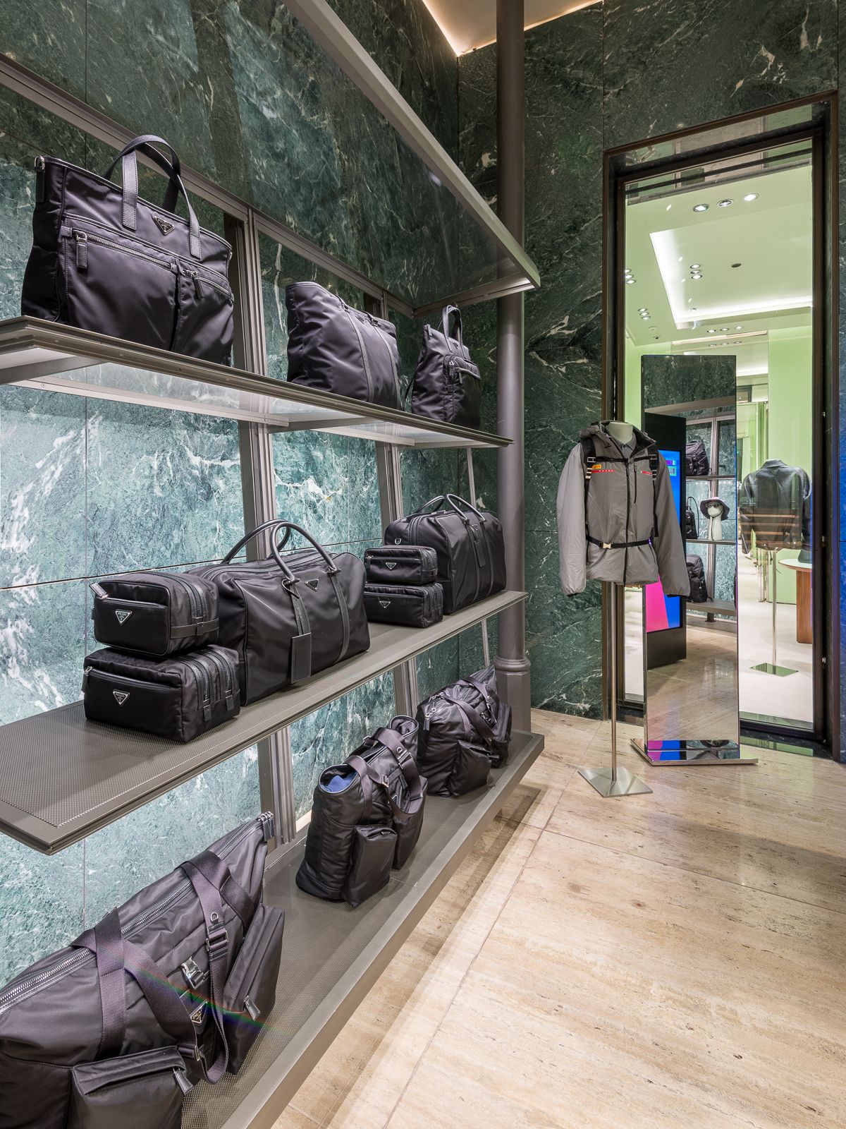 Boutique de luxe Prada amb parets de marbre verd, mostrant bosses negres de dissenyador en prestatges de metall elegant. Un maniquí amb roba grisa i una motxilla al costat d'un mirall i una obertura a una altra secció.