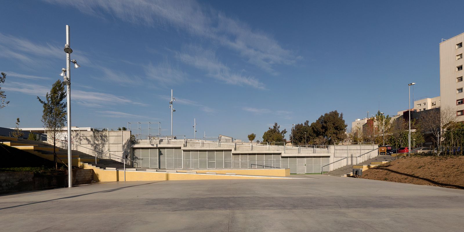 low-angle architectural photograph of Foneria Park's urban plaza, showing a large concrete foreground leading to multi-level concrete structures, yellow-accented retaining walls, metal handrails, and tall spotlights, bordered by trees and residential buildings under a blue sky.