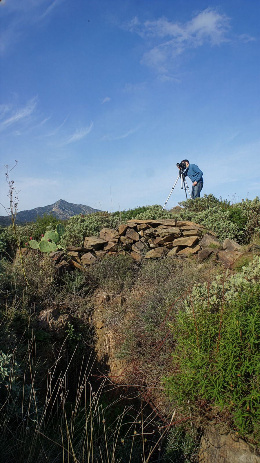 A photographer using a tripod on a rocky hillside, capturing a landscape scene with mountains in the background under a clear blue sky.