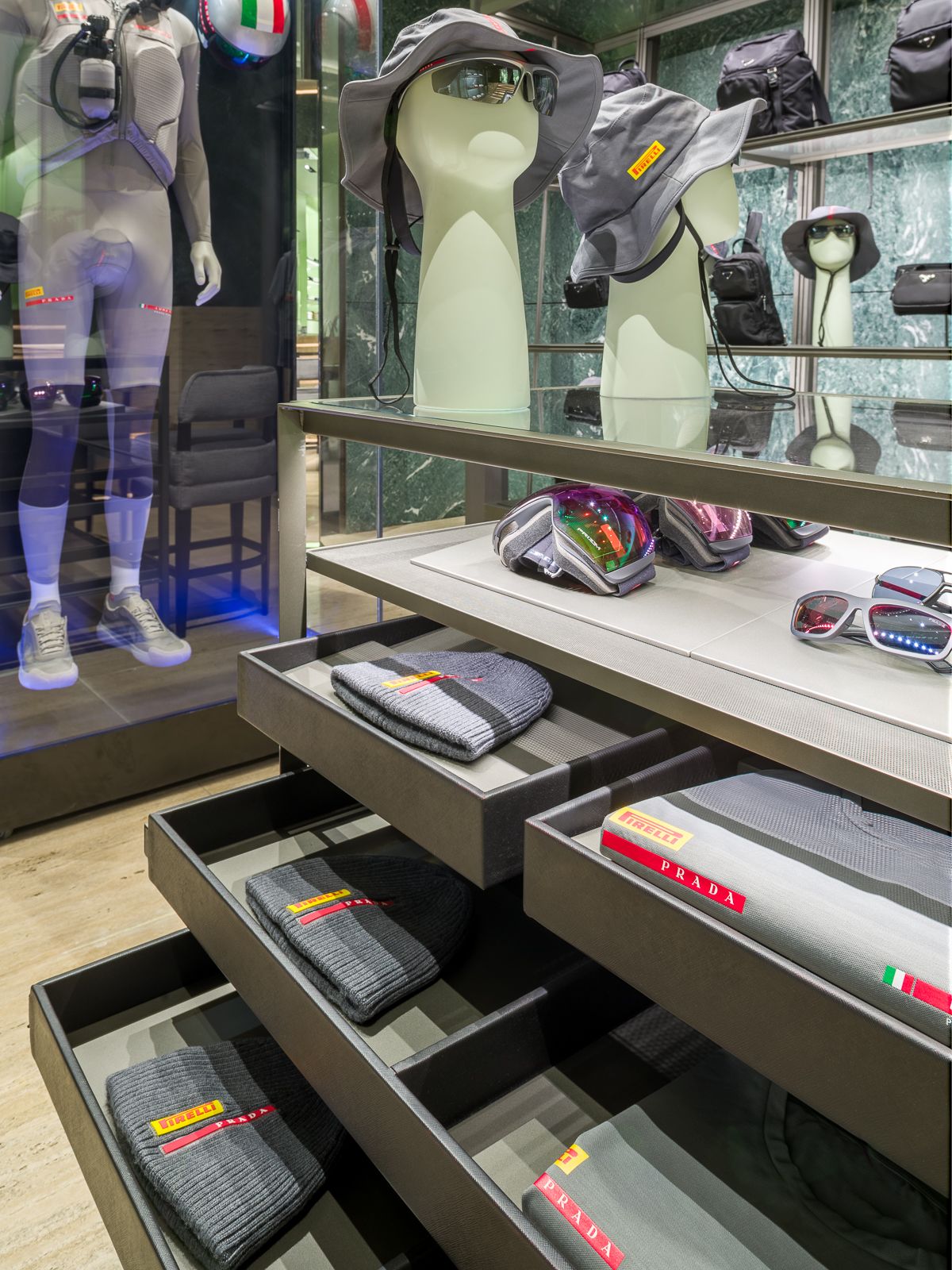 Boutique de luxe Prada, una taula de vidre amb calaixos de metall mig oberts mostrant roba grisa amb les marques Prada i Pirelli. Al fons un maniquí amb un vestit de rendiment està tancat en una vitrina de vidre.