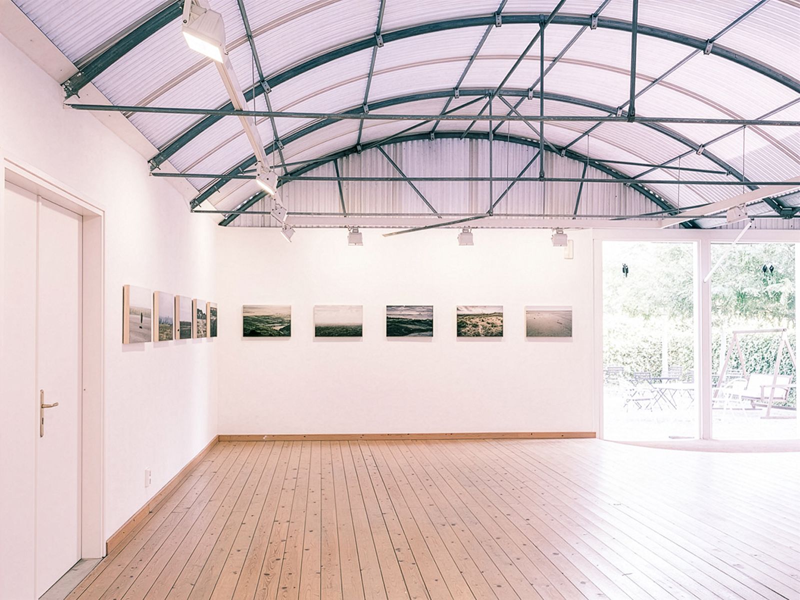 Sala de galería con techo curvo y fotografías de paisajes en las paredes. Gran puerta a la derecha abierta hacia un jardín.