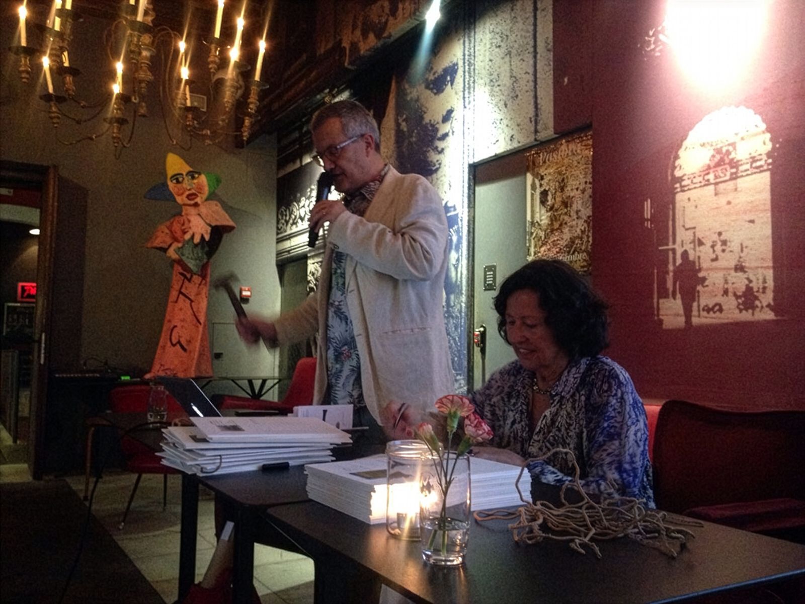 Un home parlant per un micròfon al costat d'una dona asseguda en una taula, amb decoració artística i il·luminació càlida al fons.