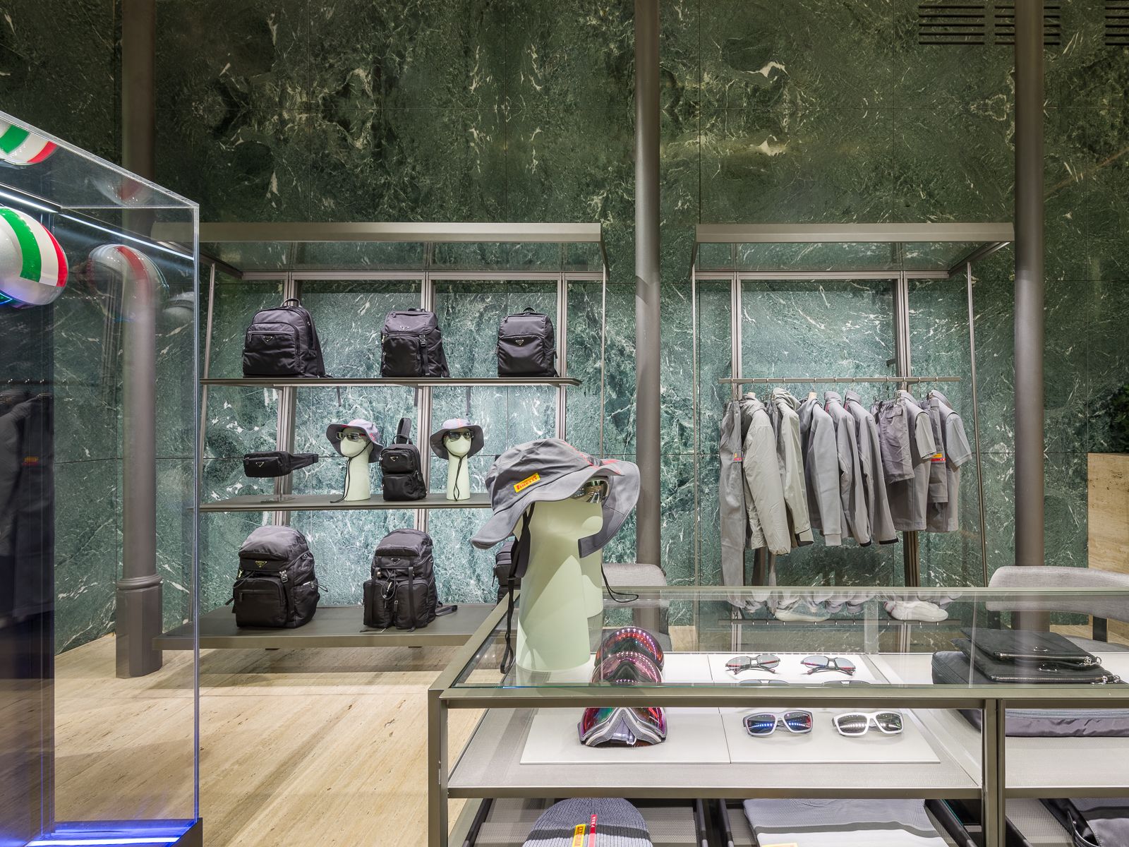 Boutique de luxe amb parets de marbre verd, mostrant sabatilles esportives de dissenyador, bosses negres i accessoris en prestatges de metall elegant. Una taula de vidre mostra ulleres i barrets, mentre que un maniquí amb un vestit de rendiment està tancat en una vitrina de vidre.