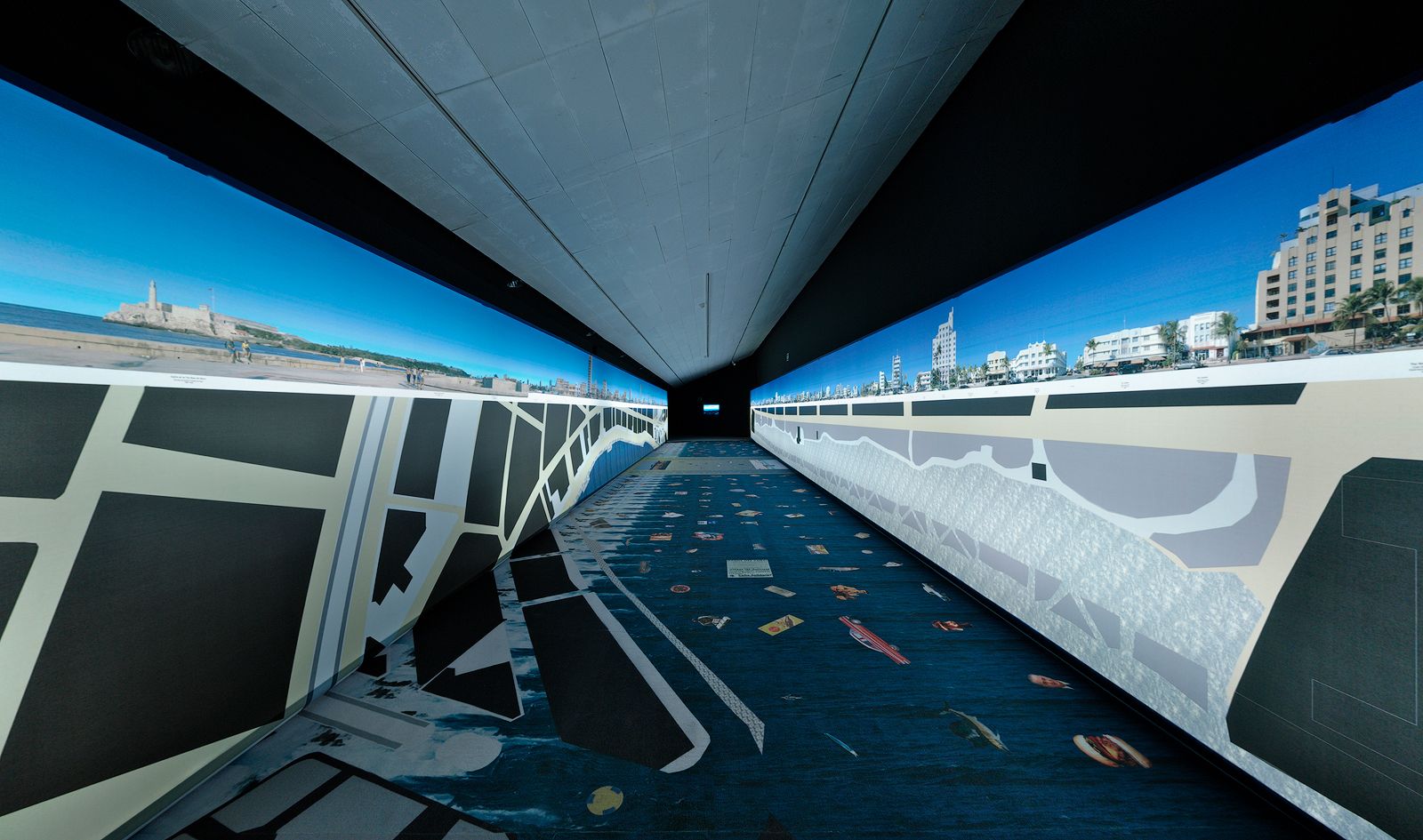 Una sala d'exposició llarga i estreta amb imatges panoràmiques d'un paisatge urbà i platja mostrades a les parets, amb plans arquitectònics detallats i mapes al terra.