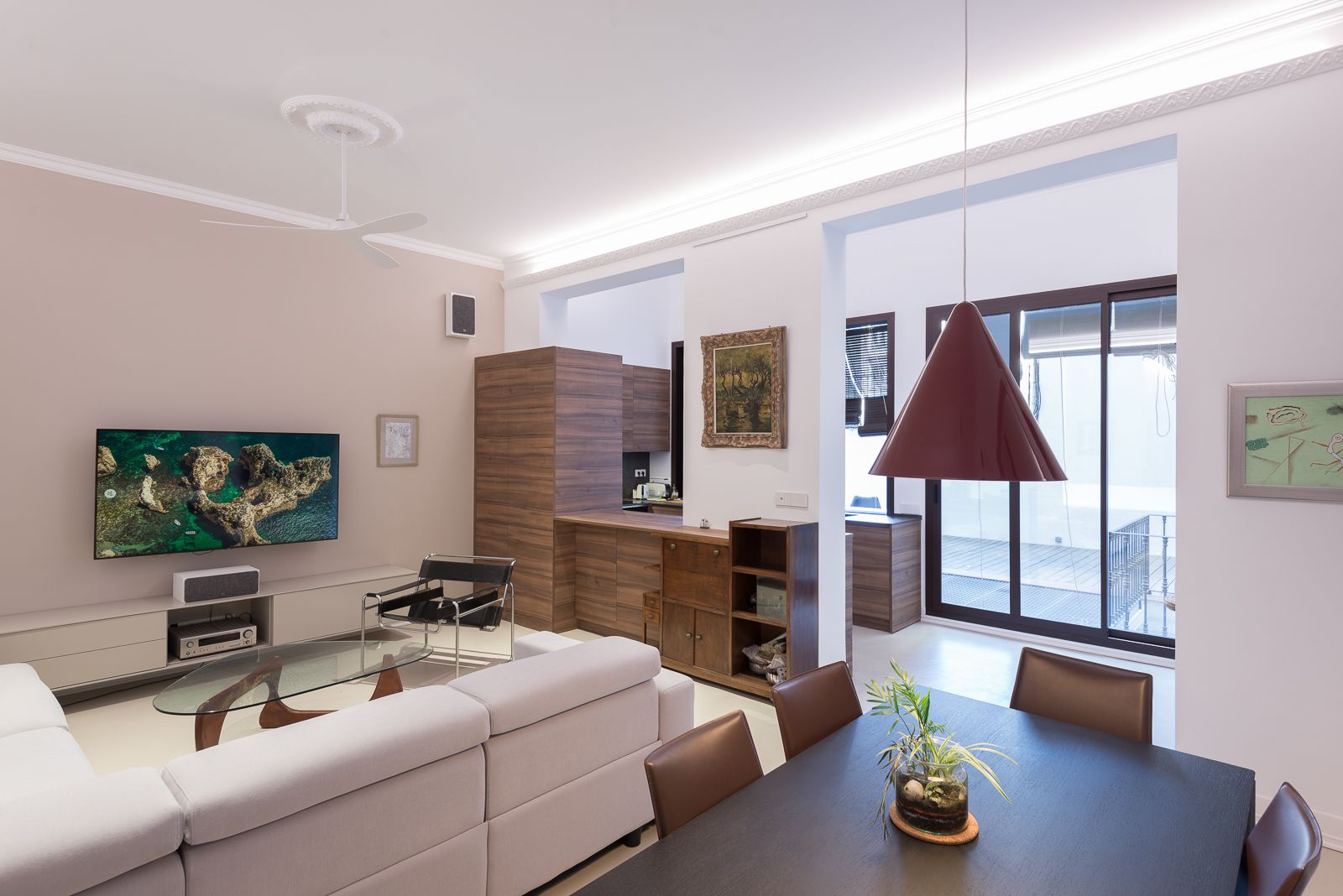 Sala d'estar moderna amb un gran televisor, sofà seccional blanc, taula de cafè de vidre i zona de menjador. Accés al pati a través de portes corredisses de vidre.