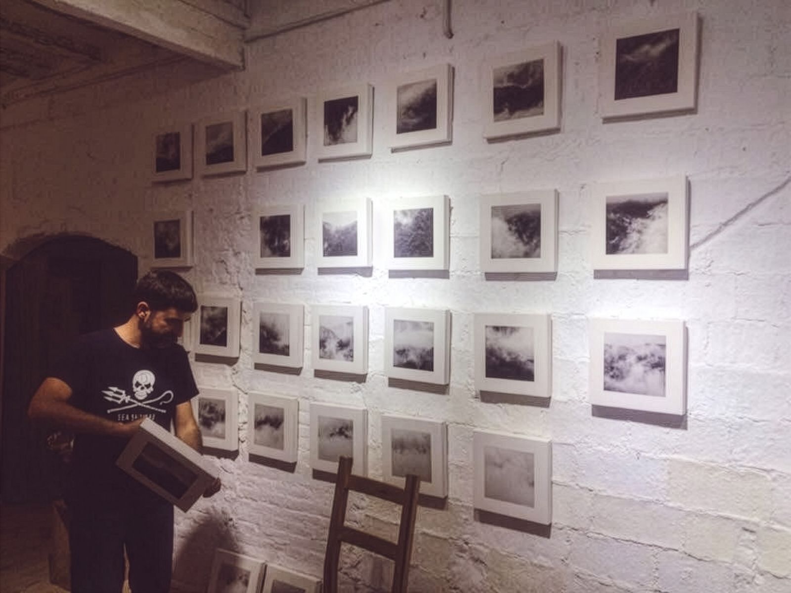 Un home col·locant fotografies en blanc i negre en una paret de totxo blanc, amb diverses ja exposades en un patró de graella, en un entorn de galeria.