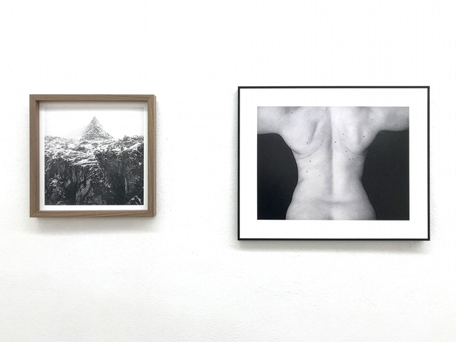 Dos fotografías enmarcadas en blanco y negro: una de un pico montañoso y otra de la espalda de una persona, expuestas en una pared blanca de galería.