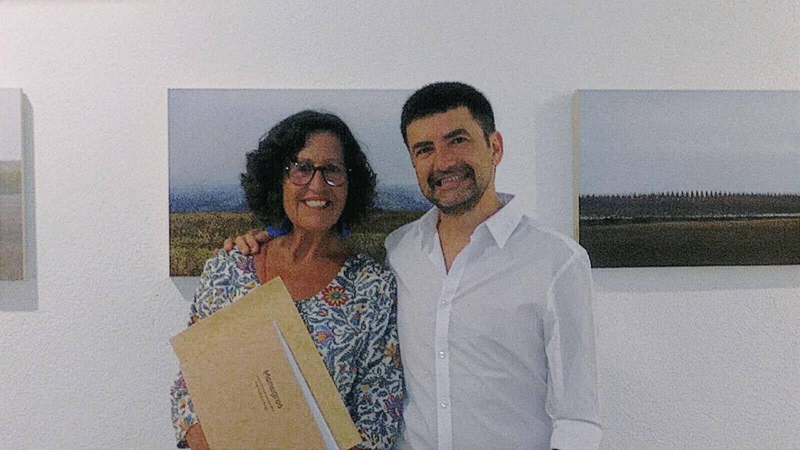 Dos personas de pie frente a una pared con fotografías de paisajes, sonriendo a la cámara, una sosteniendo un libro. El fondo muestra un paisaje tranquilo y sereno en las fotografías.