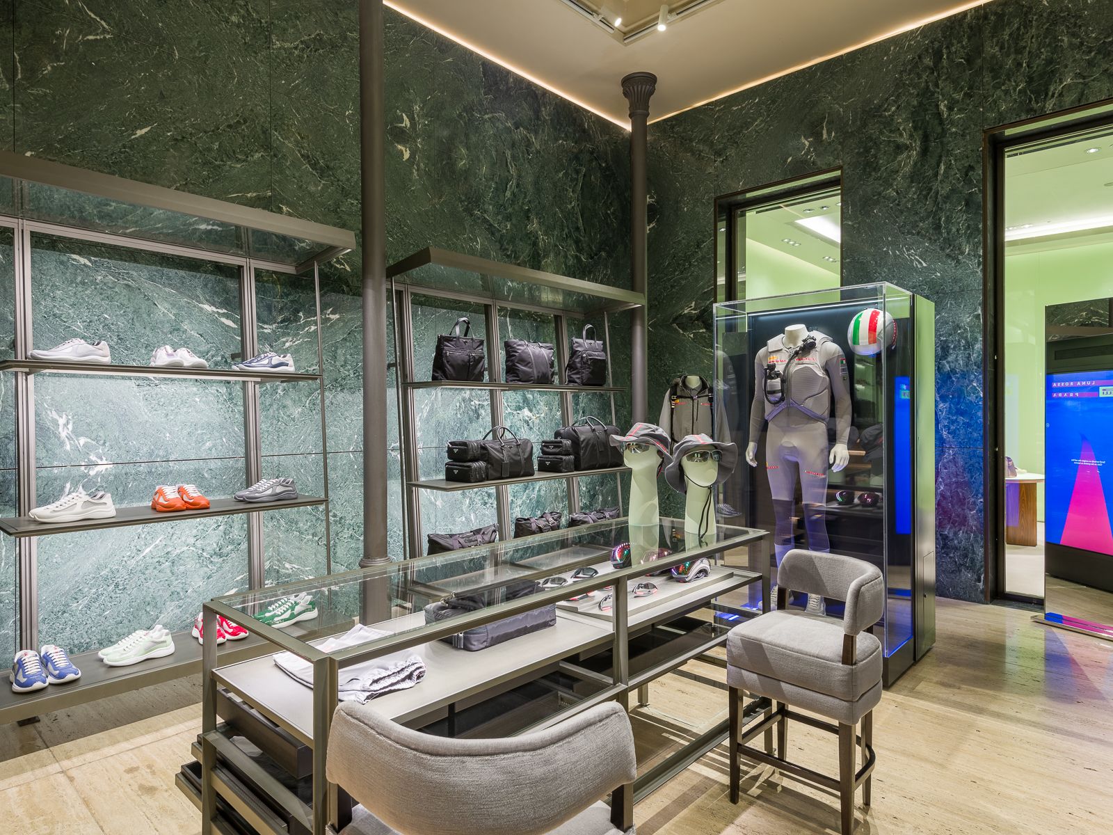 Interior d'alta gamma de boutique amb parets de marbre verd, prestatges de metall elegant mostrant sabatilles esportives i bosses, i una vitrina de vidre amb un maniquí amb un vestit de rendiment amb un casc de competició. Una taula de vidre central conté accessoris, amb seients de felpa a prop.