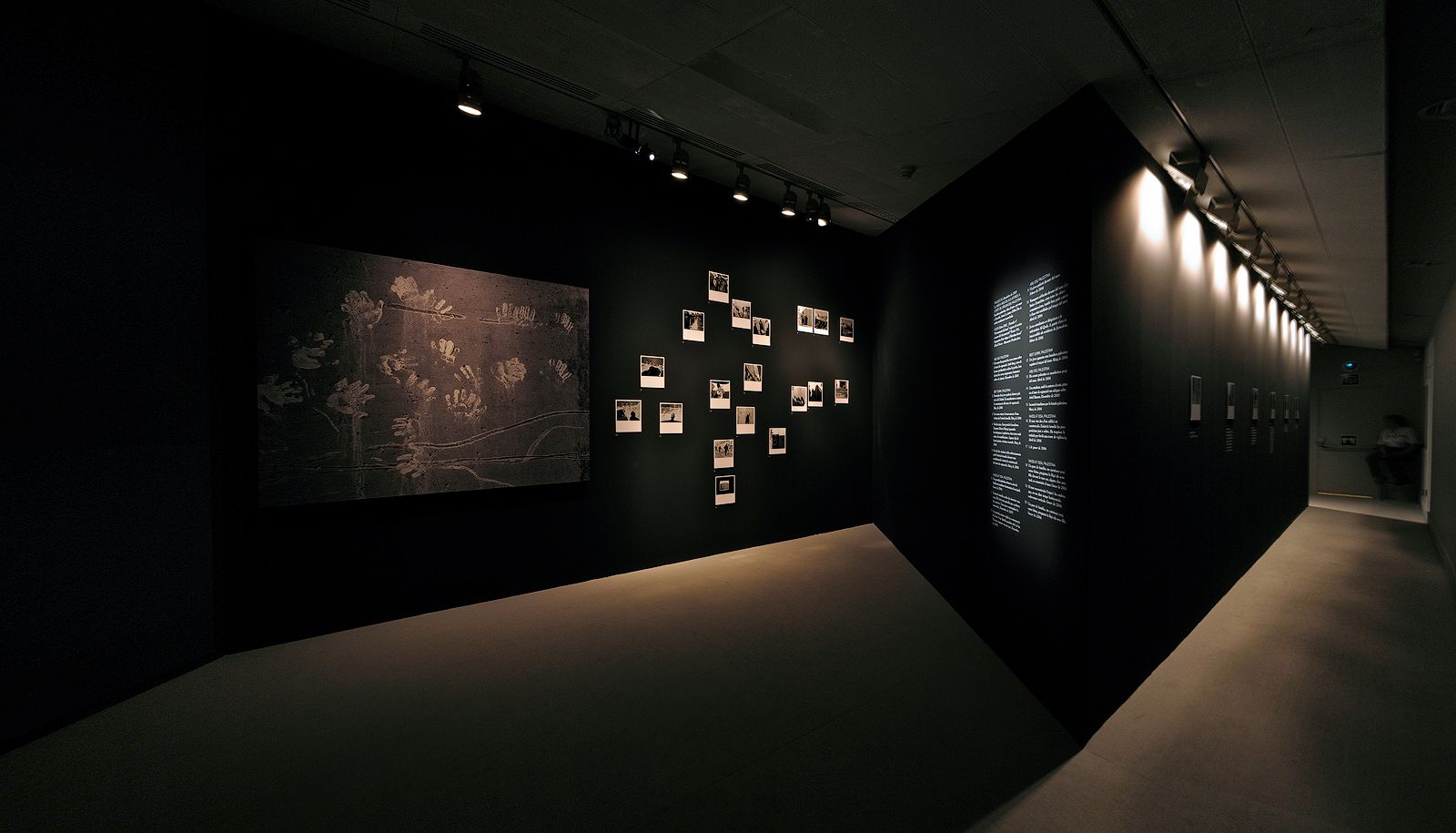 Fotografia panoràmica d'una sala d'exposicions d'art amb il·luminació tènue i parets de color negre mat, que mostra una gran obra amb empremtes de mans, un conjunt de petites fotografies i un passadís contigu il·luminat.