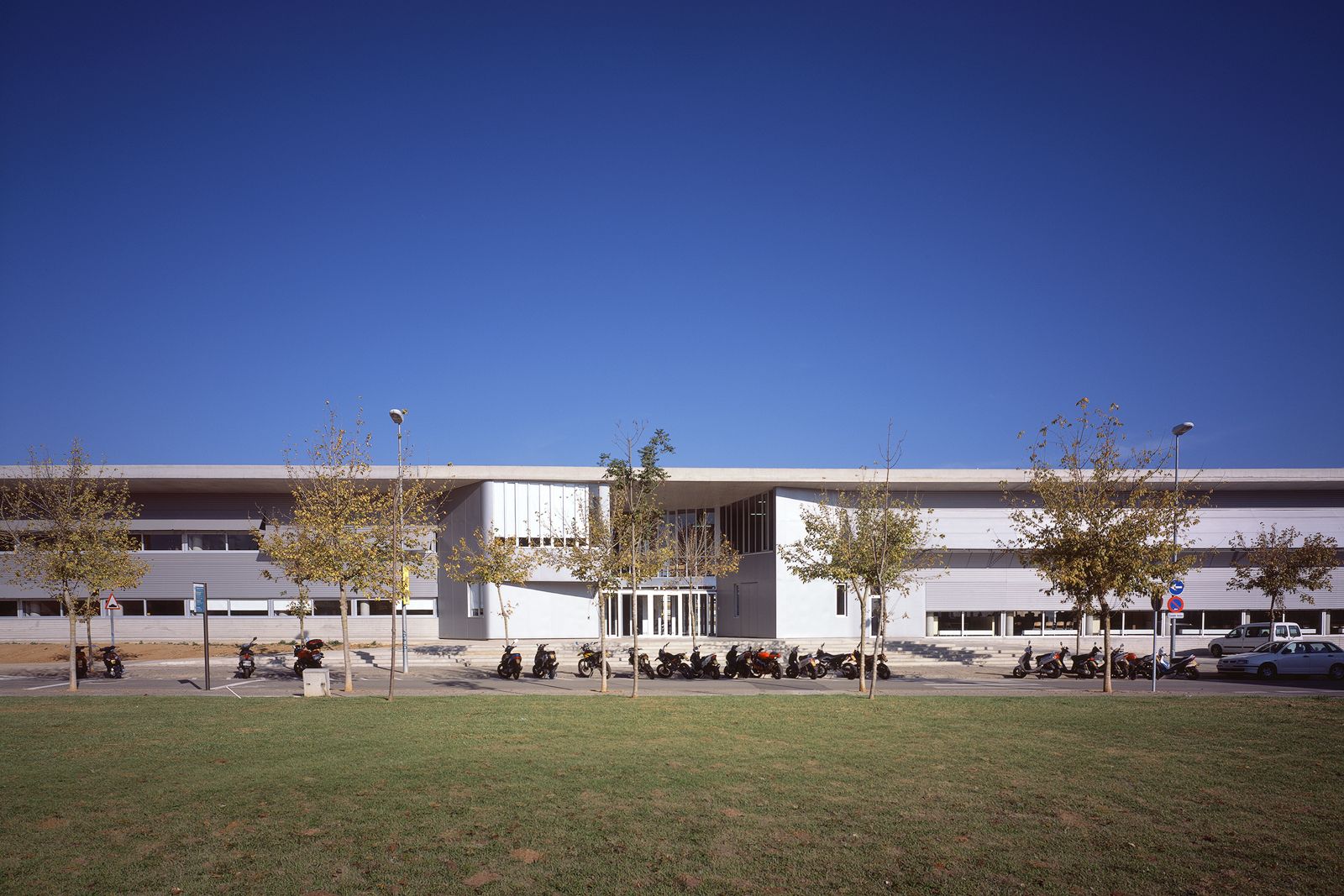 A view of Fontajau High School's modern exterior with a row of trees and parked motorcycles in front, against a clear blue sky.