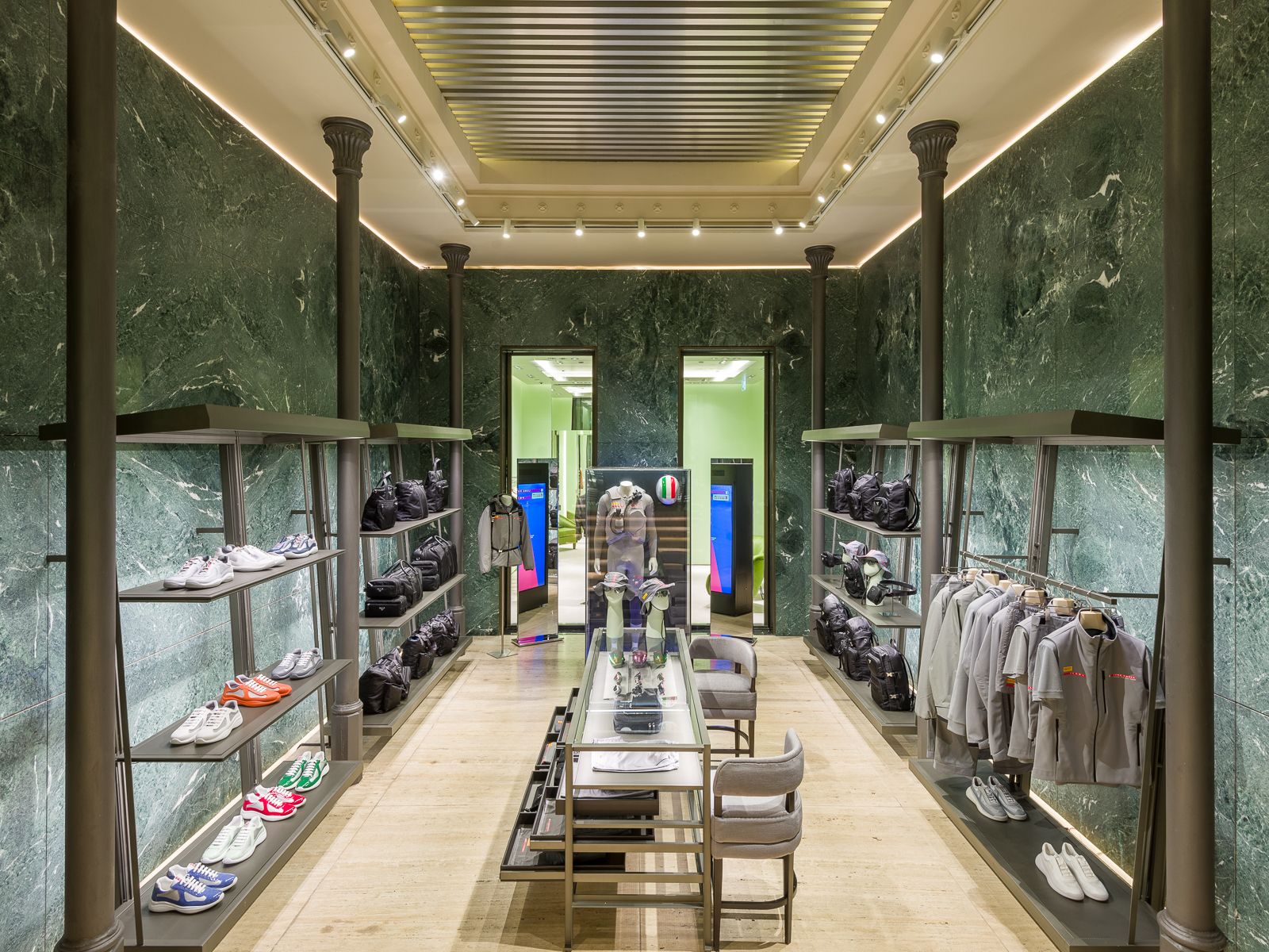 Interior luxós de boutique Prada amb parets de marbre verd, il·luminació elegant i prestatges de metall negre mostrant sabatilles esportives, bosses i roba grisa. Una taula de vidre central mostra accessoris, mentre que una porta al fons condueix a una altra secció.