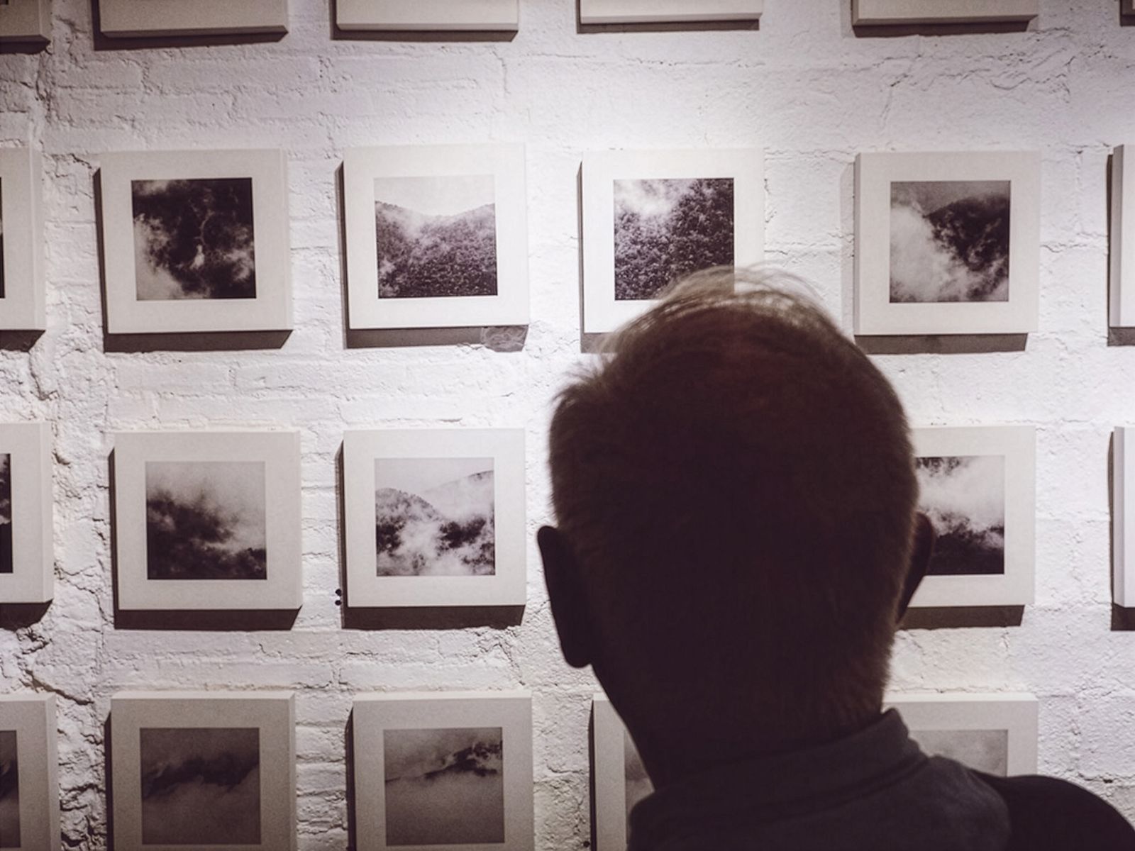 Una persona veient una gran col·lecció de fotografies quadrades en blanc i negre de núvols i boira en una paret de totxo blanc.