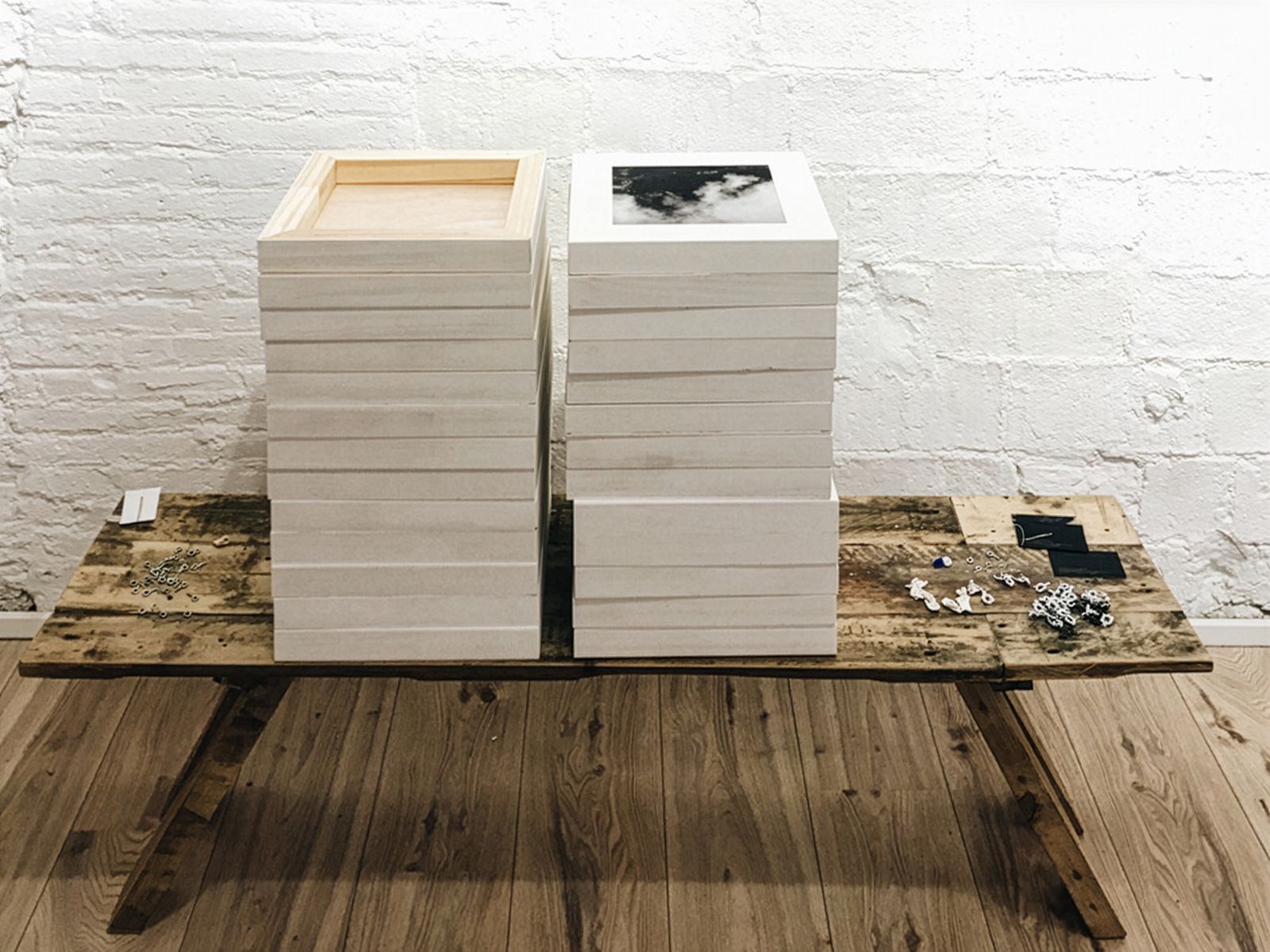 Dues piles de fotografies emmarcades en blanc sobre una taula de fusta, al costat de petits ganxos de metall i altres materials de penjat, en una galeria amb parets de totxo blanc.