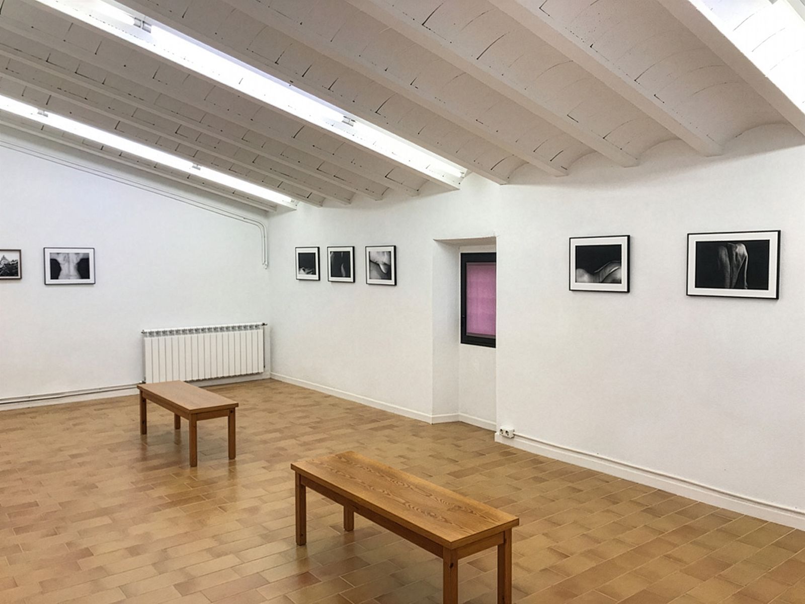 Una sala de galería minimalista con fotografías en blanco y negro en paredes blancas, dos bancos de madera en el centro y una pequeña ventana en una pared.