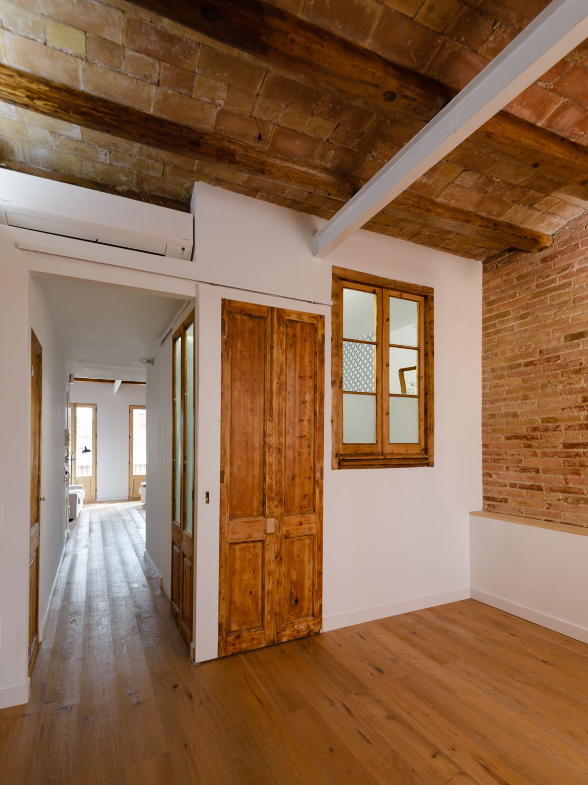 Hallway communicating two living areas, with wooden doors and windows, exposed brick walls, and wooden beams.