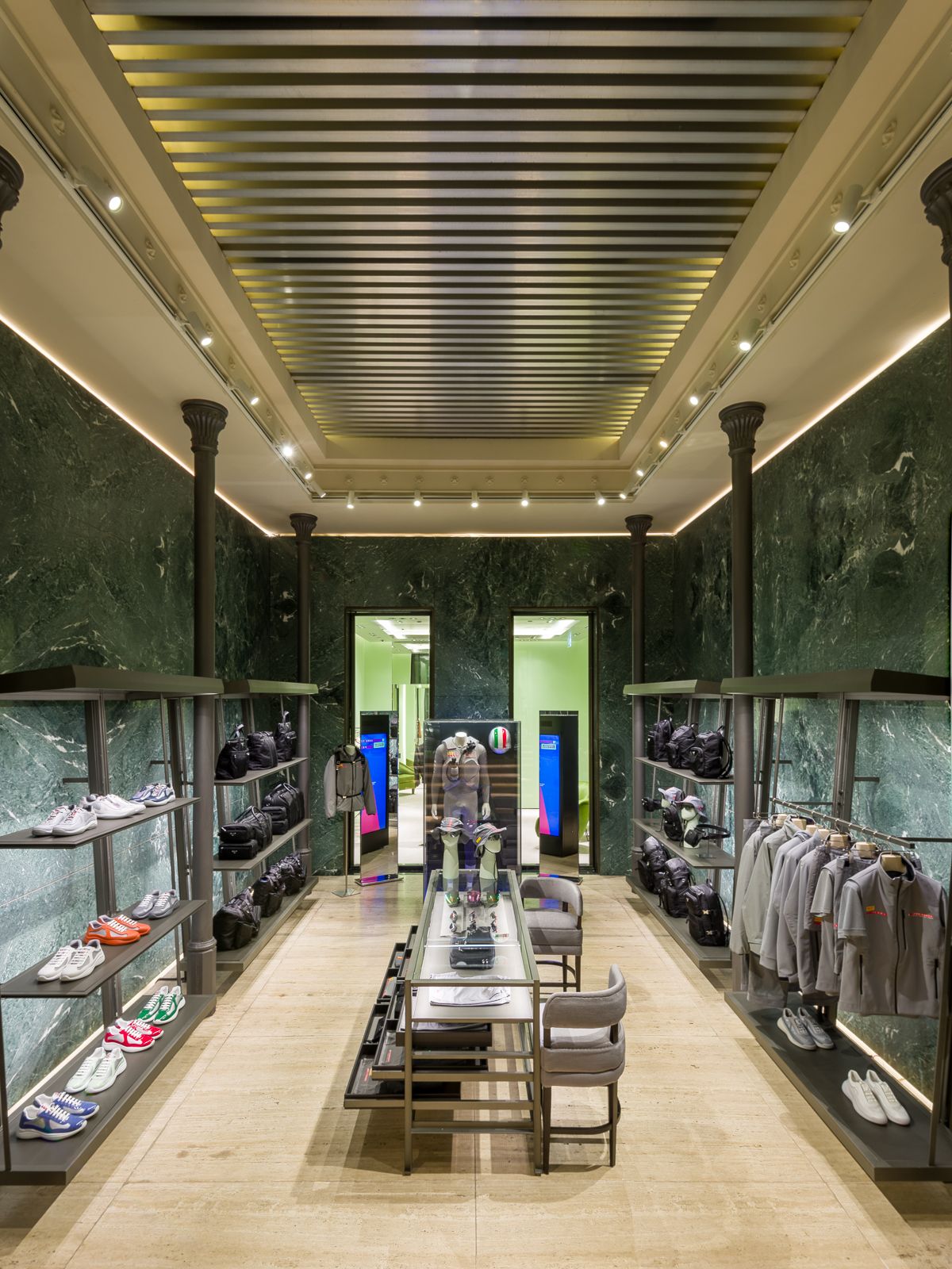 Interior luxós de boutique amb parets de marbre verd, il·luminació elegant i prestatges de metall negre mostrant sabatilles esportives, bosses i roba grisa. Una taula de vidre central mostra accessoris, mentre que una porta al fons condueix a una altra secció.