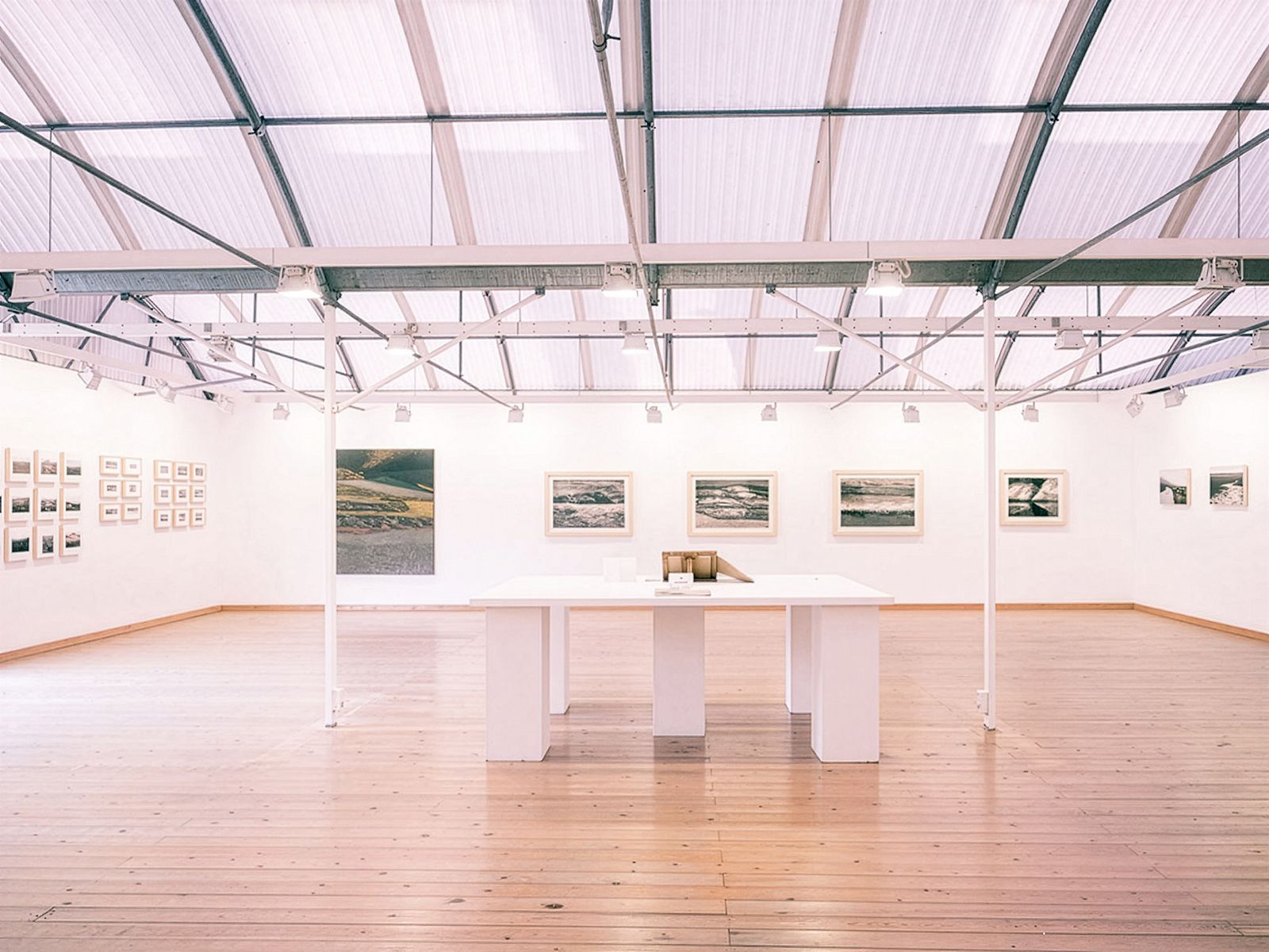 Sala de exposición luminosa con obras de arte enmarcadas en las paredes, un techo curvo alto, suelos de madera y una mesa blanca central con libros expuestos.