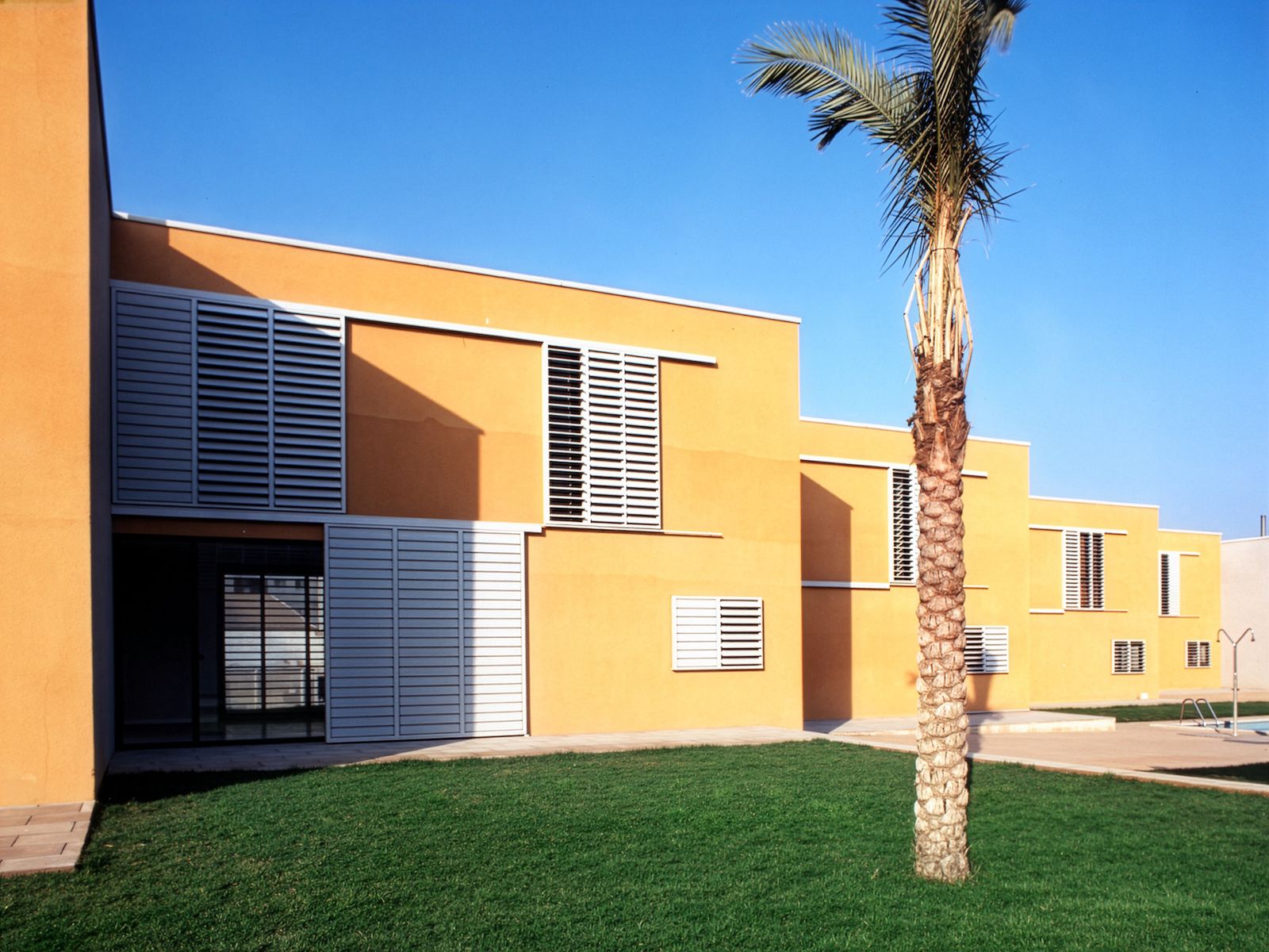 A cluster of modern, flat-roofed buildings with yellow facades and large louvered windows, set against a bright blue sky and featuring a palm tree in the foreground.