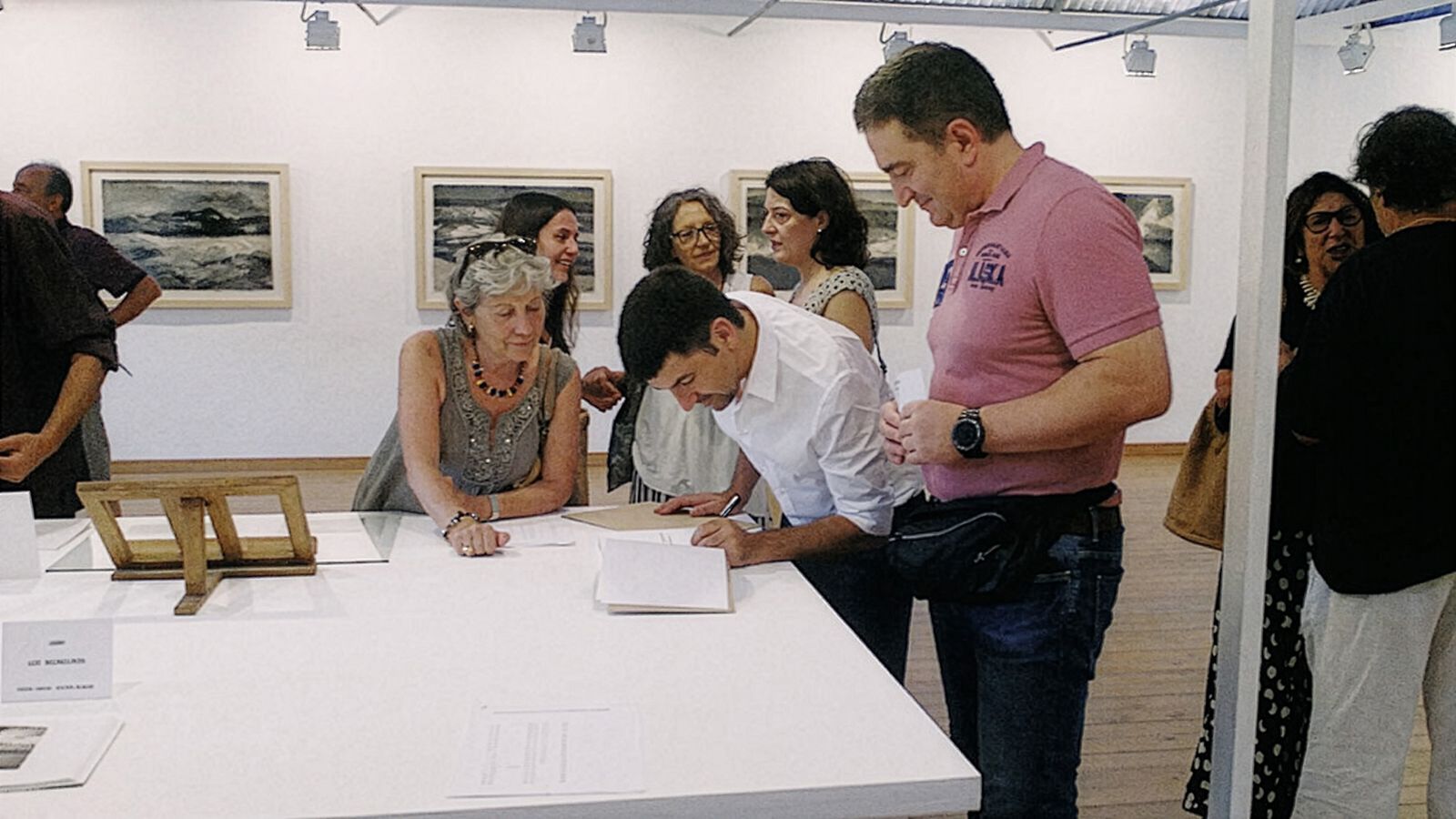 Personas reunidas en una galería de arte, interactuando con el arte y entre ellas. Una persona está firmando un libro en una mesa, mientras otros están conversando y viendo fotografías de paisajes expuestas en las paredes al fondo. La atmósfera es animada e interactiva.