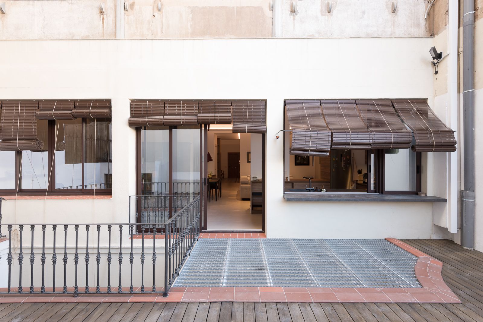 Pati exterior amb barana de metall, coberta de fusta i grans finestres amb persianes de bambú, que condueixen a un interior modern.