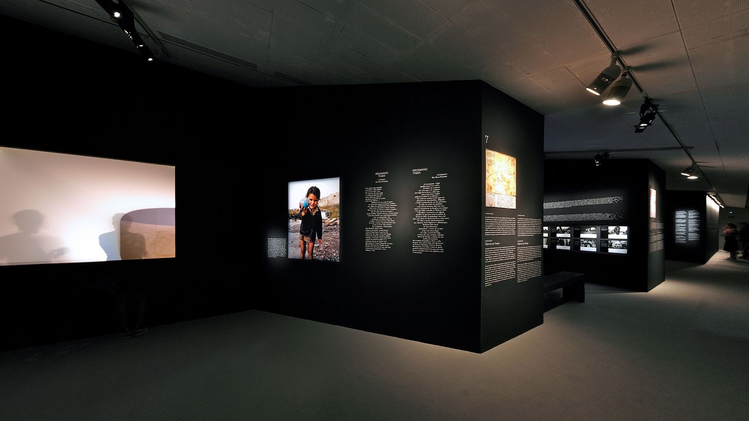 Vista d'angle ampli d'una sala d'exposicions d'un museu modern amb plafons expositius en negre mat amb blocs de text, una fotografia il·luminada d'un nen, una pantalla de projecció brillant a l'esquerra i un recorregut que continua al fons.