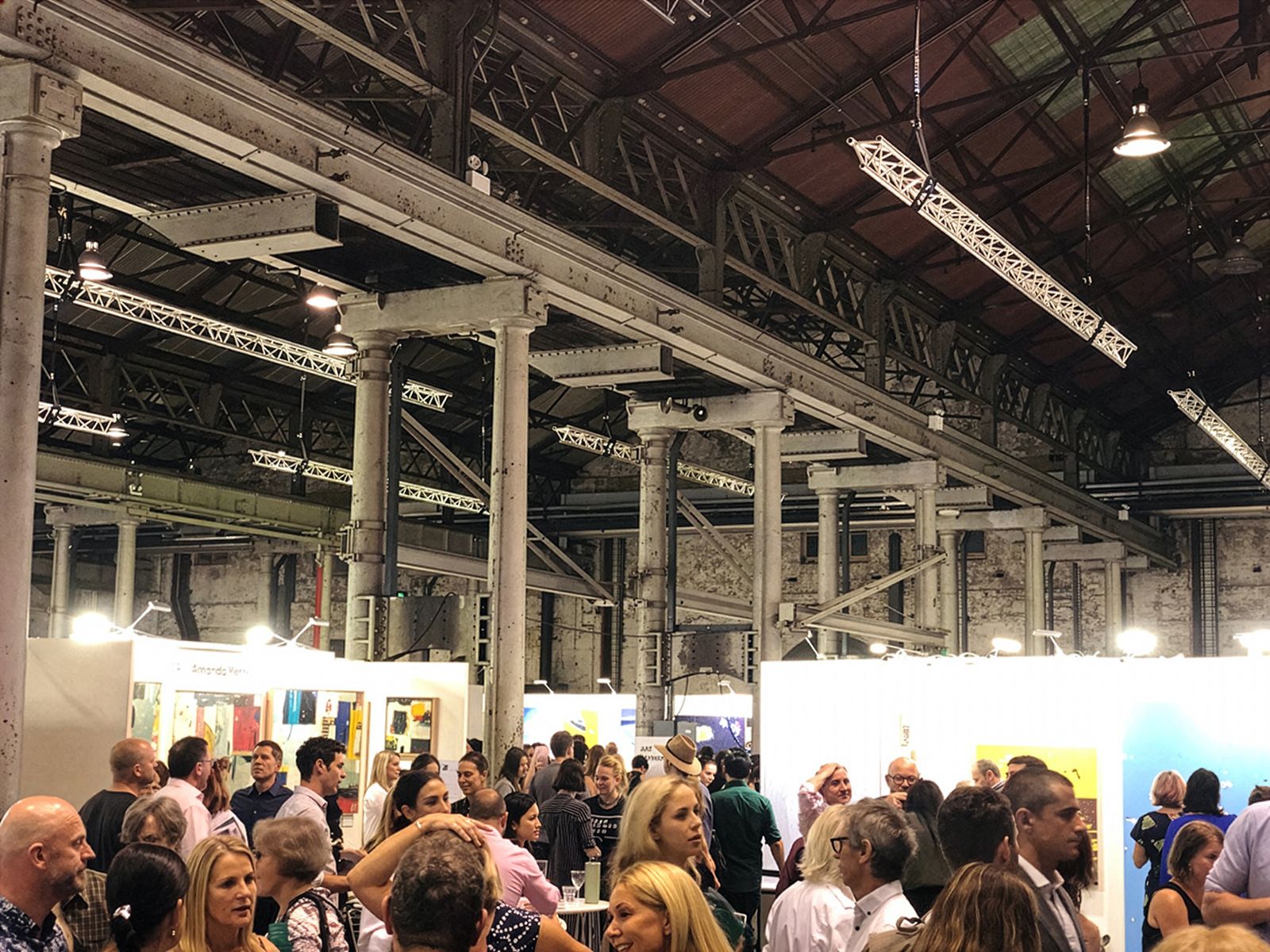Una exposició d'art plena de gent amb moltes persones veient diverses obres d'art en un espai ampli d'estil industrial amb sostres alts i bigues exposades.